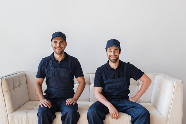joyful workmen in overalls and caps smiling at camera while sitting on white couch - Zdjęcie, obraz