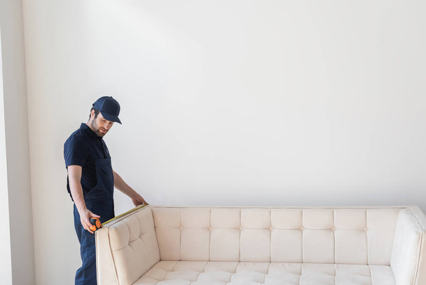 young mover in cap and overalls measuring couch near white wall - Foto, Imagen