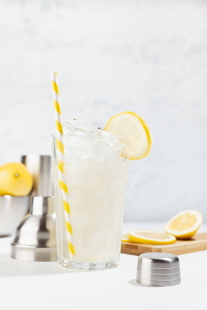 Cooking lemon juice in soft light fresh elegant white kitchen in sunny day - lemonade in misted glass with straw, ice cubes with ingredients, silver bowl, shaker, vertical. - Photo, Image