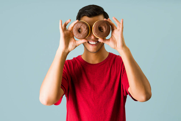 Happy young man smiling while holding two chocolate donuts in front of his eyes and having fun - 写真・画像