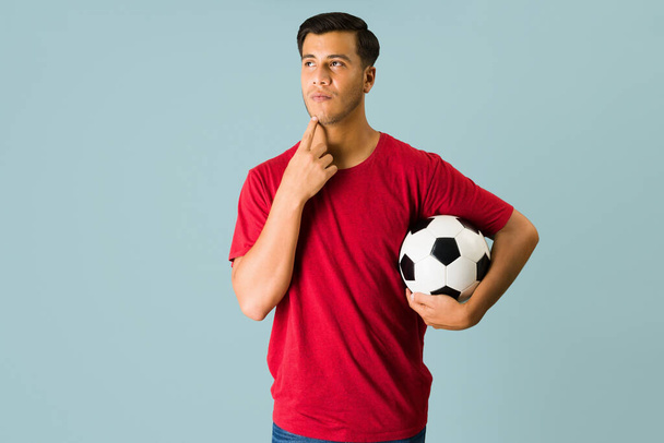 Спортивный латиноамериканец думает о том, чтобы пойти поиграть в футбол, держа мяч под мышкой. - Фото, изображение
