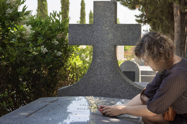 Jonge vrouw, met hoofd gebogen en verdrietig knielend naast een marmeren graf met een groot kruis en een kruis gegraveerd, waardoor een takje bloemen op een graf. Op de achtergrond zijn cipressen uit beeld. - Foto, afbeelding