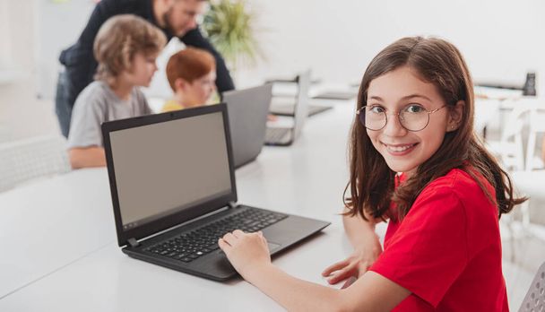 Cheerful schoolgirl with laptop in classroom - Фото, изображение