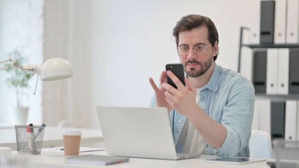 Young Man with Laptop using Smartphone at Work - Photo, Image