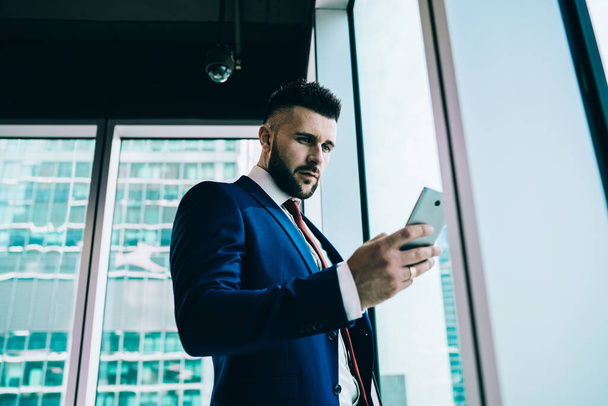 From below elegant confident executive man focusing on screen and interacting with smartphone while standing against window of high rise office building in daytime - Zdjęcie, obraz