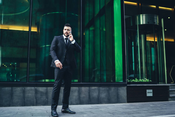 Modern male boss in black suit and tie discussing business problem by mobile phone while standing next to contemporary office center  - Фото, изображение
