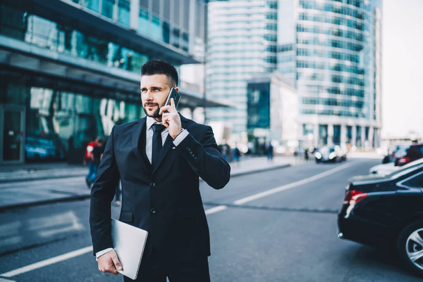 Líder masculino positivo con tableta mirando hacia otro lado y contestando una llamada telefónica mientras está parado en la carretera en el centro de la ciudad moderna - Foto, imagen