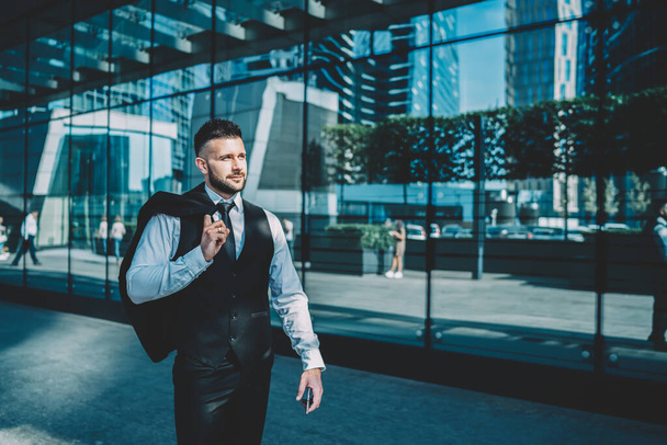 Confident contented executive man in formal clothes with jacket over shoulder looking away and enjoying life while walking alone after work along street against glass facade with reflection of skyscrapers in sunny day - Foto, Bild