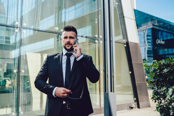 Serious bearded male in suit adjusting jacket and frowning while looking away and answering phone call outside contemporary building in city - Φωτογραφία, εικόνα