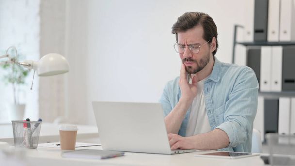 Hombre joven con dolor de muelas trabajando en el ordenador portátil - Foto, Imagen
