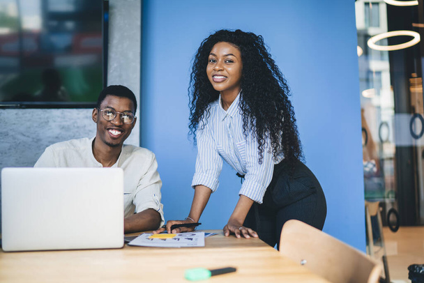Delighted African American partners in modern formal clothes surfing laptop and studying documents at contemporary workplace and looking at camera - Photo, Image