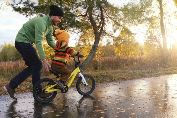 Father teaches his little child to ride bicycle in autumn park. Happy family moments. Time together dad and son. Candid lifestyle photo. - Foto, Imagem