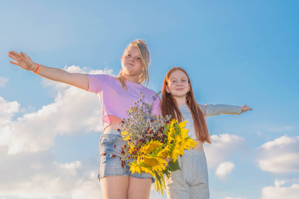Dvě mladé dívky v letních šatech stojí proti modré obloze. Jedna paže byla natažená na bok jako křídla letadla. Ostatní ruce drží kytici žlutých slunečnic a divokých květin. Podívejte se do kamery. - Fotografie, Obrázek