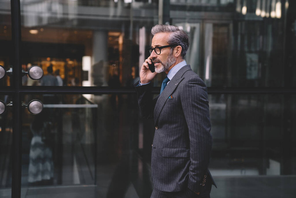 Vista laterale dell'uomo d'affari con la barba in elegante abito scuro che parla al telefono vicino alla parete di vetro dell'edificio per uffici mettendo mano in tasca a New York - Foto, immagini