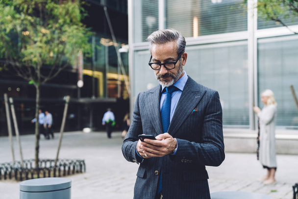 Middelbare leeftijd zakenman in bril en stijlvolle donker gestreepte business suit staan in de stad straat en browsen smartphone in handen in New York - Foto, afbeelding