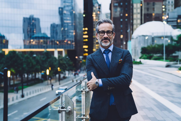 Handsome adult man in glasses and business suit with arms crossed grinning and standing on background of evening New York street while looking at camera - Valokuva, kuva
