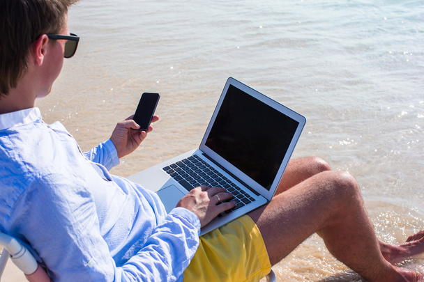 Young businessman using laptop and telephone on tropical beach - Photo, Image