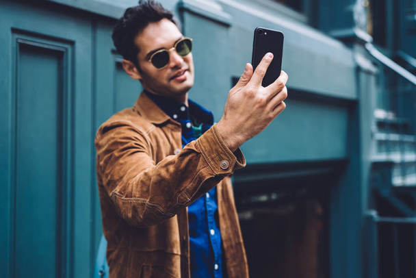 Young handsome man in trendy clothes and sunglasses taking selfie on smartphone while standing against blue building of New York street - Fotografie, Obrázek