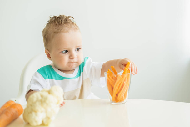 Baby boy sitting in a Childs chair eating carrot slices on white background - Foto, afbeelding
