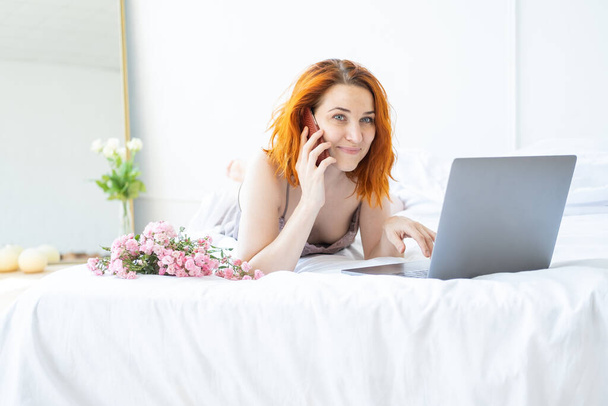 attraente zenzero donna mezza età in pigiama sdraiato a letto con telefono e laptop. - Foto, immagini