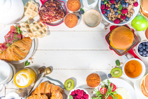 Various traditional breakfast food - fried eggs with bacon, muesli, oats, waffles, pancakes, burger, croissants, fruit berry, coffee, tea and orange juice, white table background copy space top view - Photo, Image
