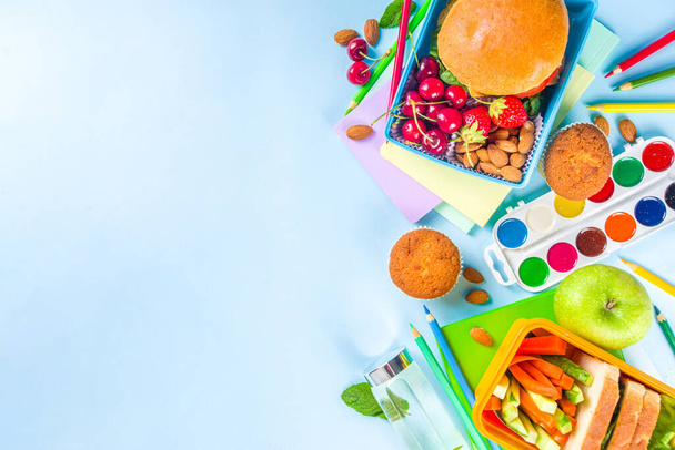 Επιστροφή στο σχολείο, Υγιεινό γευστικό παιδικό κουτί με σάντουιτς, ξηρούς καρπούς, φρέσκα φρούτα και ξυλάκια λαχανικών. Με σχολικά είδη, μολύβια, σημειωματάρια, σε φωτεινό μπλε φόντο επίπεδη κορυφή άποψη αντίγραφο χώρο - Φωτογραφία, εικόνα