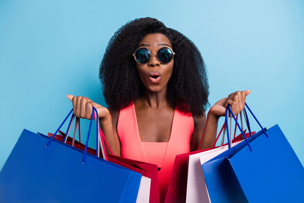 Photo portrait of curly woman surprised shocked keeping packages on sale in sunglass isolated vivid blue color background - Foto, imagen