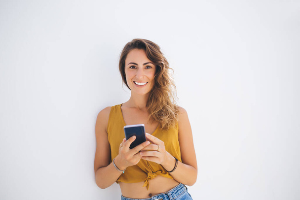 Optimistic charming female millennial in crop top leaning against white wall and smiling while texting on smartphone and looking at camera - Fotó, kép