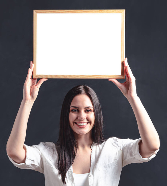 Улыбающаяся женщина держит пустой рекламный щит. Счастливая девушка в белой рубашке с плакатом в рамке над головой - Фото, изображение