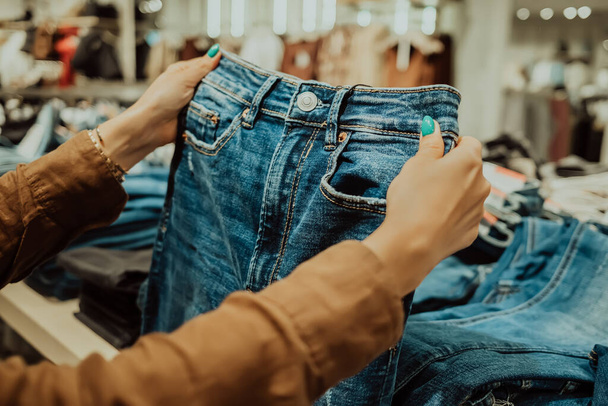 Young woman in fashion store chooses things for herself - jeans, denim trousers. Customers, shopping, sales concept. - Foto, afbeelding