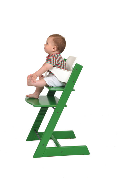 vista lateral de un retrato completo de un bebé sentado en una trona sobre fondo blanco - Foto, Imagen