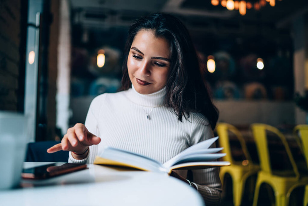 Sensual young optimistic dark haired woman in casual wear reading literature with pleasure spending time sitting at table in cafeteria - Zdjęcie, obraz