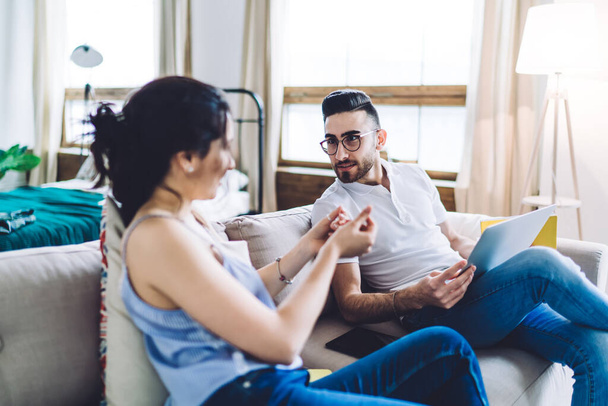 Young ethnic man with bristle and glasses in casual clothes and with laptop listening attentively and looking at woman while sitting together on sofa at home - Foto, afbeelding