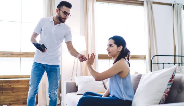 Side view of tense woman sitting on sofa and showing by hand refuse of credit card that smart bearded man suggesting standing in living room - Foto, immagini
