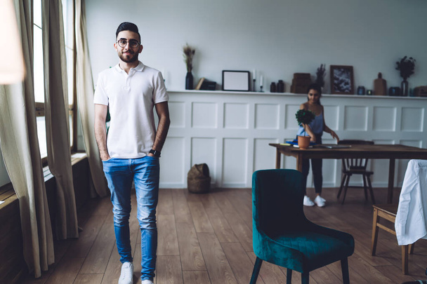 Young modern male standing next to velvet chair and keeping hands in jeans pockets while woman organizing table in background at home - Foto, immagini