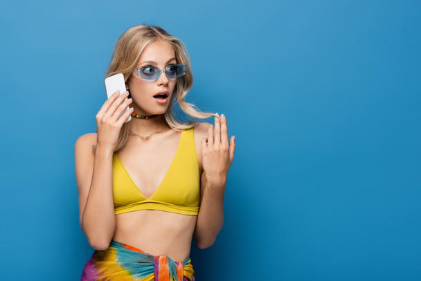 shocked young woman in yellow bikini top talking on smartphone isolated on blue - Photo, image