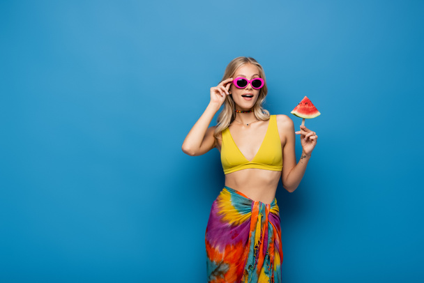 amazed young woman adjusting sunglasses and holding popsicle stick with watermelon on blue - Fotoğraf, Görsel