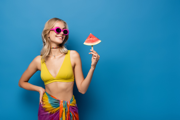 glückliche junge Frau, die mit der Hand auf der Hüfte steht und Eis am Stiel mit Wassermelone auf blau hält - Foto, Bild