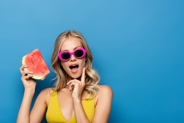 surprised young woman in pink sunglasses holding slice of watermelon isolated on blue - Φωτογραφία, εικόνα