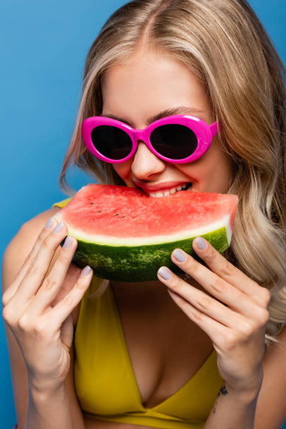 fröhliche junge Frau mit pinkfarbener Sonnenbrille isst frische Wassermelone isoliert auf blauem Grund - Foto, Bild
