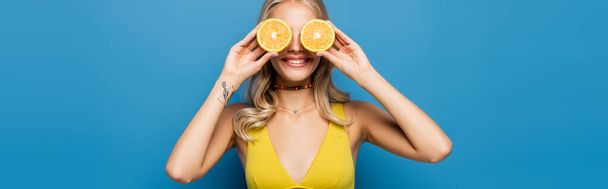 cheerful young woman in yellow bikini top covering eyes with orange halves isolated on blue, banner - Fotografie, Obrázek