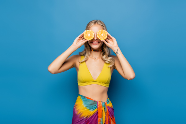 alegre joven en bikini amarillo cubriendo los ojos con mitades anaranjadas en azul - Foto, Imagen