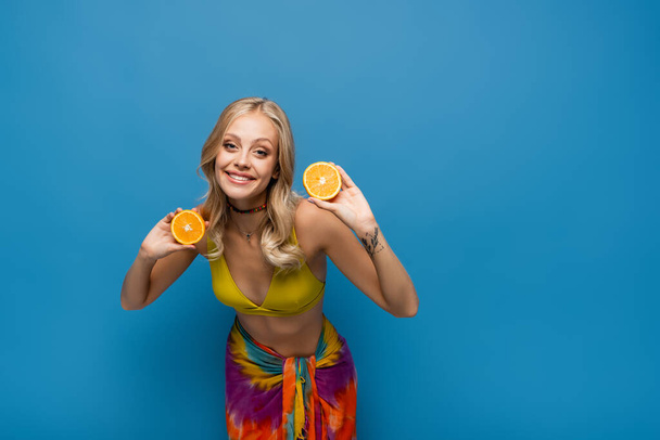smiling young woman in yellow bikini top holding orange halves on blue - Photo, Image