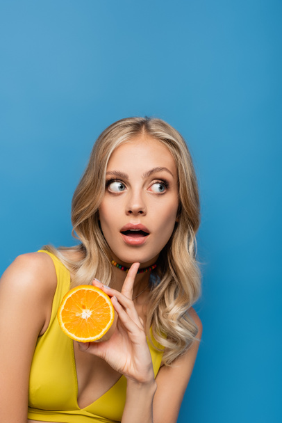 surprised young woman in yellow bikini top holding orange half isolated on blue - Fotografie, Obrázek