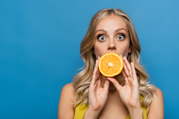 šokovaná mladá žena zakrývající ústa s oranžovou polovinou izolované na modré - Fotografie, Obrázek