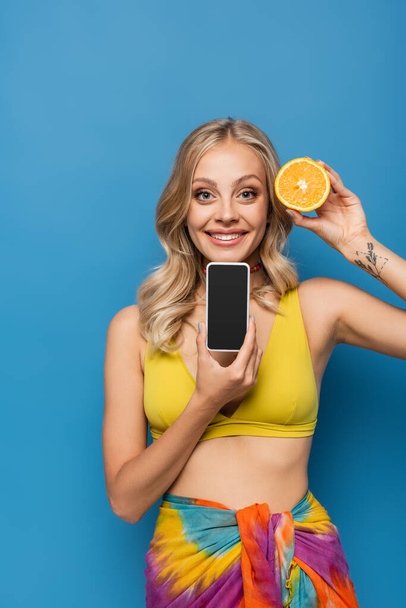 幸せな若いです女性で黄色ビキニトップ保持オレンジ半分とスマートフォンとともに空白の画面で隔離された青 - 写真・画像