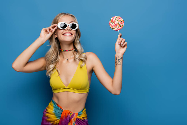 smiling young woman in yellow bikini top and sunglasses holding sweet lollipop on blue - 写真・画像