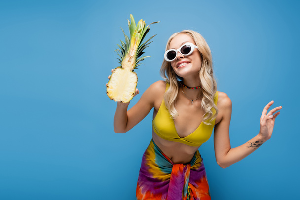 smiling young woman in sunglasses and bikini top holding sweet pineapple half isolated on blue - Фото, изображение