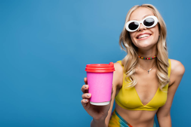 happy young woman in sunglasses and bikini top holding paper cup isolated on blue  - Фото, зображення
