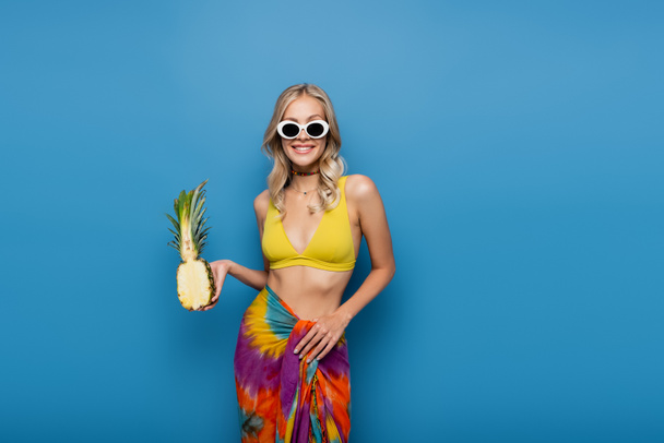 radostná žena v slunečních brýlích a bikinách top drží sladký ananas napůl izolované na modré - Fotografie, Obrázek